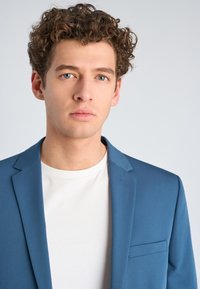 Giovane uomo con capelli ricci castani e occhi azzurri che indossa un blazer blu sopra una camicia bianca, espressione neutra, sfondo grigio chiaro.