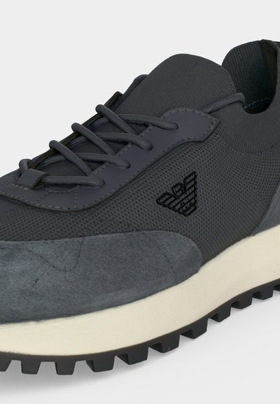Baskets gris foncé avec tissu texturé et détails en daim, logo noir sur le côté, semelle blanche et semelle extérieure noire.