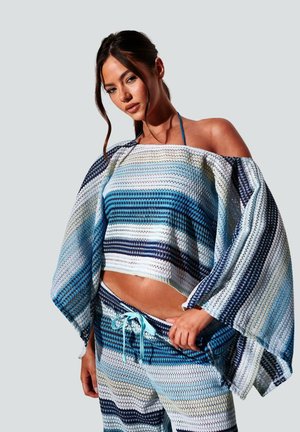 Kobieta o ciemnych włosach nosi luźny, pasiasty niebiesko-biały top z opuszczonymi ramionami oraz pasujące do niego spodnie, pozuje na jednolitym tle.