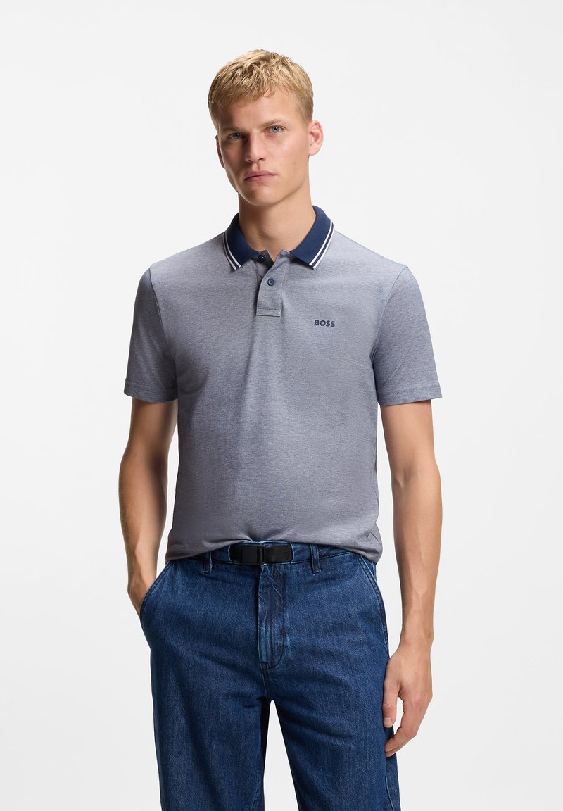 Νέος άνδρας με κοντά ξανθά μαλλιά, φοράει γκρι πόλο πουκάμισο BOSS με σκούρο μπλε γιακά και μπλε τζιν, στέκεται με τα χέρια στις τσέπες.