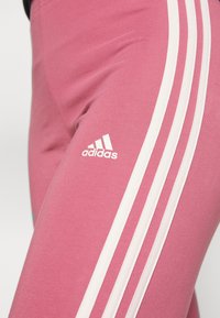 Pink athletic leggings featuring three vertical white stripes on the side and a small white Adidas logo near the hip. Smooth texture.