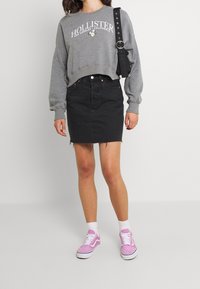 Sweat-shirt gris court avec logo blanc et accents floraux, associé à une jupe en jean noire. Le modèle porte des baskets en daim rose avec des semelles blanches.