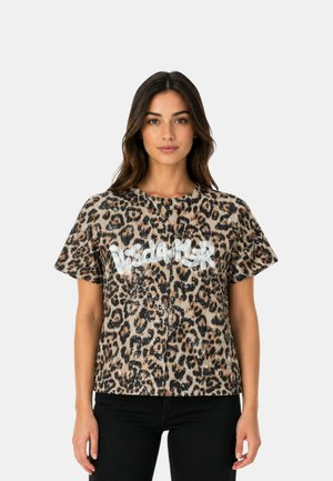 Donna che indossa una camicia a maniche corte con stampa leopardata con la parola "Disclaimer" in lettere bianche, abbinata a pantaloni neri, in piedi su uno sfondo semplice.