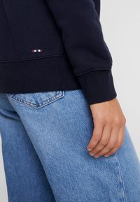 Sudadera navy con puños de canalé combinada con jeans de mezclilla azul claro. La sudadera presenta un bordado sutil cerca del dobladillo. Ajuste casual y relajado.