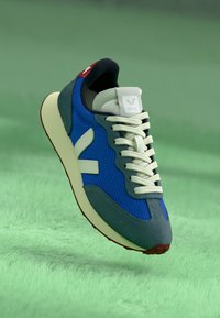 Blå sneakers laget av mesh og semsket skinn har en hvit logo, grå detaljer og beige snøringer. Sålen er kremfarget med en rød hældetalj.