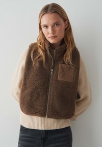 Gilet en polaire marron avec fermeture éclair à l'avant et col haut, doté d'une poche plaquée beige. Porté par-dessus un pull léger, sur un fond neutre.