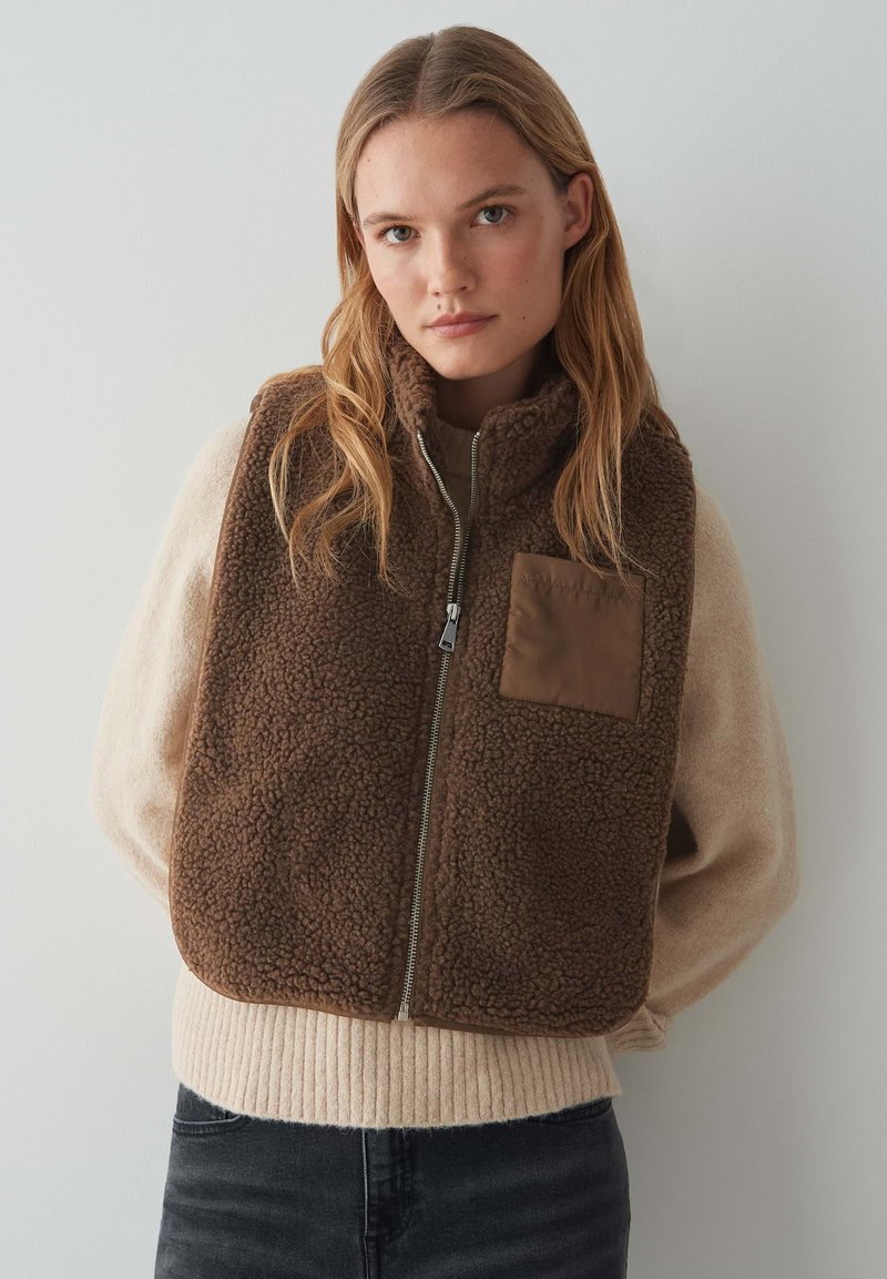 Gilet en polaire marron avec fermeture éclair à l'avant et col haut, doté d'une poche plaquée beige. Porté par-dessus un pull léger, sur un fond neutre.