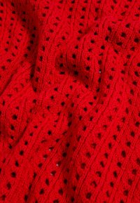Tissu tricoté rouge vif avec des rangées alternées de petits trous carrés et des motifs en torsades texturés, présenté en plis doux.