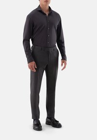 Camicia lunga grigia scuro con colletto, abbinata a pantaloni grigi scuro strutturati e scarpe eleganti nere, in piedi su uno sfondo semplice.