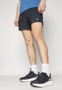 Schwarze Sportshorts mit seitlichem Schlitzdesign und gesticktem Logo. Kombiniert mit leichten schwarz-weißen Laufschuhen und weißen Socken.