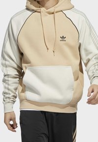 Beige hoodie med en vit framficka, svarta detaljer på ärmarna och en broderad logotyp på bröstet. Justerbara dragsko ingår.