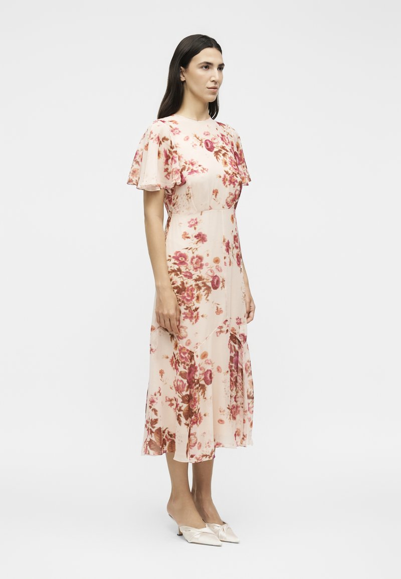 Frau steht und trägt ein hellrosa Midikleid mit rotem Blumenmuster, flatternden Ärmeln und spitzen, cremefarbenen High Heels.