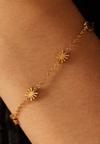 Gouden armband met afwisselende bloemenmotieven en schakelkettinkje, met gladde oppervlakken en een gepolijste afwerking, tegen een huidskleurige achtergrond.