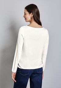 Haut à manches longues et col rond en tissu blanc doux, avec une coupe décontractée et des poignets froncés, associé à un pantalon en denim bleu foncé.