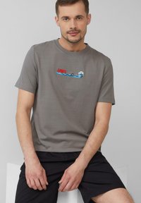 Szara bawełniana koszulka z grafiką "SZCZĘK" przedstawiającą rekina i fale w odcieniach niebieskiego i białego. Krótkie rękawy i okrągły dekolt.