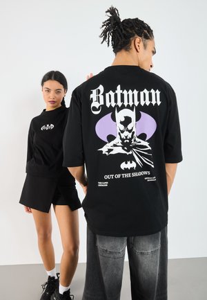 Black t-shirt featuring a graphic of Batman with purple accents and white text. The model wears a matching black outfit with shorts.