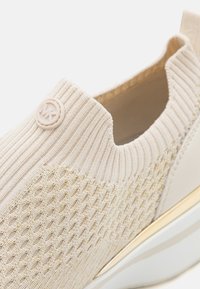 Beige strikket sneakers med teksturert design og gullakenter, en myk sokklignende krage og en sirkulær logo på siden.