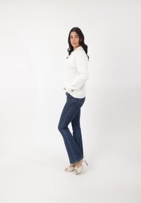 Suéter blanco de canalé, jeans acampanados azules y tacones abiertos con patrones y acentos metálicos. La modelo está de lado contra un fondo blanco.