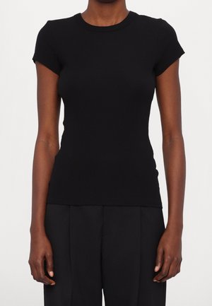 Vrouw draagt een nauwsluitend zwart T-shirt met korte mouwen en ronde hals en losse zwarte broek, staand met ontspannen armen langs het lichaam.