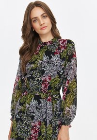 Floral dress in sheer black fabric, featuring green, pink, and red botanical patterns. High neck, long sleeves with elastic cuffs, fitted waist.