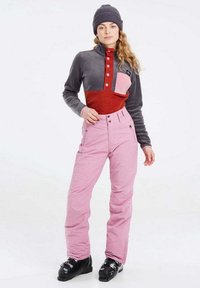 Fleece pulóver s farebnými blokmi v šedej, červenej a ružovej farbe; ružové lyžiarske nohavice; čierne lyžiarske topánky; model má na sebe pletenú čiapku.