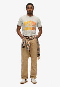 Grå t-shirt med orange bjerggrafik, beige bukser og en ternet skjorte bundet om taljen. Hvide sneakers med sorte accenter.