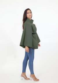 Grön cape-stil jacka med hög krage och framsidan knappring, matchad med blå skinny jeans och klara klacksandaler.