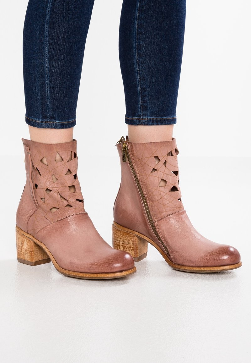 Bottes en cuir rose poussiéreux montant jusqu'à la cheville, avec talons blocs en bois, fermetures éclair latérales et motifs découpés géométriques, portées avec un jean skinny bleu foncé.