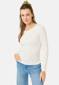 Top bianco a maniche lunghe con scollo tondo e dettagli laterali sottili, abbinato a jeans in denim blu chiaro. Vestibilità classica.