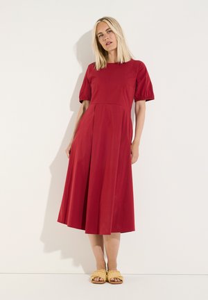 Woman with straight blonde hair wears a red midi dress with puff sleeves and yellow woven slip-on sandals, standing against a plain wall.