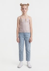 Young girl with blonde hair in two buns and braids, wearing a pink striped tank top, light blue jeans, white socks, and white sneakers, standing.