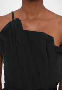 Black one-shoulder top with pleated texture and thin strap, showcasing a smooth fabric surface and clean lines.