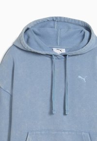 Šviesiai mėlynas džemperis su laisvu kirpimu, turintis priekinę kišenę, raištelius ir mažą Puma logotipą ant krūtinės. Minkšta tekstūra.