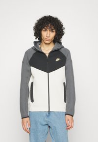 Hoodie Nike con un design a blocchi di colore in bianco, grigio e nero, con zip frontale, tasche laterali e logo sul petto.