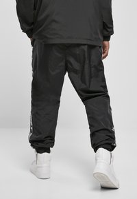 Schwarze, strukturierte Windbreaker-Hosen mit elastischen Bündchen und seitlichen Logo-Akzenten, kombiniert mit weißen High-Top-Sneakern.
