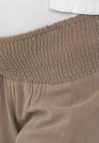 Gros plan d’un pantalon beige avec une large ceinture élastiquée smockée portée sur un haut blanc côtelé.