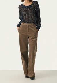 Hihnainen tummansininen paita, jossa on valkoisia pilkkuja, yhdistetty korkeavyötäröisiin, leveälahkeisiin ruskeisiin corduroy-housuihin. Mustat kärkitossut.