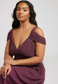 Dramatisk violett klänning med axelsnitt, texturerad tyg och rynkade detaljer. Modell med långt hår, sittande med armarna korsade.