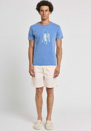 Homme debout portant un T-shirt bleu avec un motif porte-clés, un short beige clair et des chaussures beiges faciles à enfiler sur un fond clair uni.