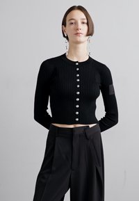 Zwarte geribbelde cardigan met lange mouwen, ronde hals en grote zilveren knopen. Gecombineerd met high-waisted zwarte broek.