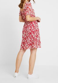 Rotes Blumenkleid mit kurzen Puffärmeln, einem taillierten Oberteil und ausgestelltem Rock, kombiniert mit weißen Turnschuhen. Strukturierter Stoff mit weißen Blumen.