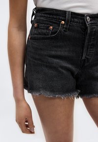  Κοντινό πλάνο ενός ατόμου που φοράει ψηλόμεσα μαύρα τζιν σορτς με ξεφτισμένα τελειώματα και χαλκινούς κοχλίες, συνδυασμένα με λευκή μπλούζα.