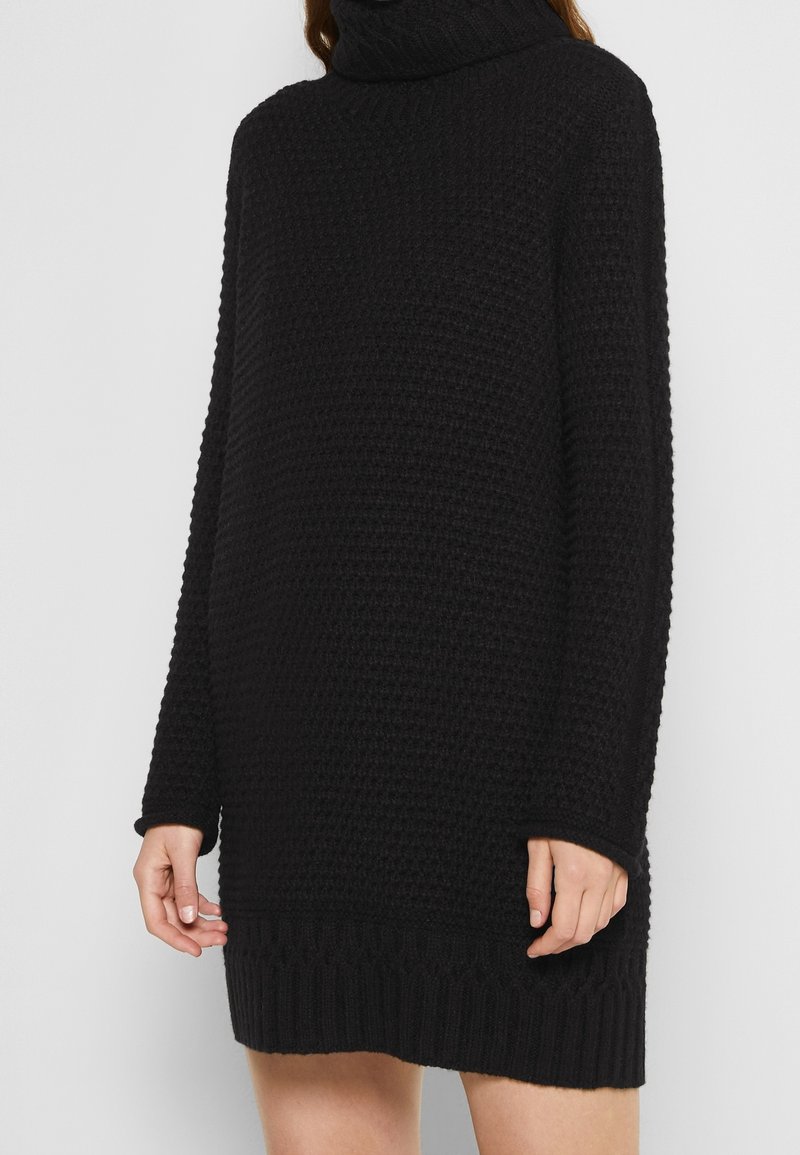 Femme portant une longue robe pull en tricot noir à col roulé avec des manches longues sur un fond uni.