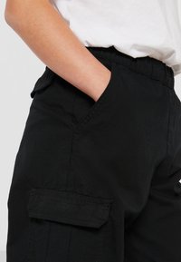 Pantalons cargo noirs en tissu texturé, avec une taille élastique et des poches latérales. Présente un détail de poche à rabat sur la jambe.