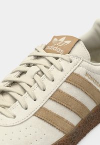Ελαφρώς μπεζ αθλητικό παπούτσι Adidas με τρεις καφέ σουέτ ρίγες, ταμπά ετικέτα με το λογότυπο στη γλώσσα και υφασμένη καφέ λαστιχένια σόλα.