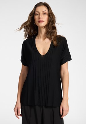 Kvinde med bølget brunt hår iført en sort t-shirt med korte ærmer og V-hals med lodrette folder og sorte bukser mod en ensfarvet baggrund.