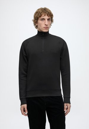 Jeune homme aux cheveux bouclés portant un pull noir à manches longues avec un col en demi-fermeture éclair et un logo discret sur la poitrine, debout devant un fond uni.