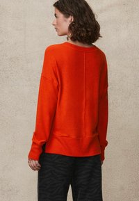 Helles oranges Sweatshirt mit lockerer Passform, gerippten Bündchen und Saum sowie einer Mittelnähten auf der Rückseite. Getragen mit gemusterten schwarzen Hosen.