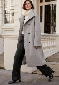 Cappotto grigio di lana con colletto a revers, tre bottoni neri, tasche laterali, indossato sopra un dolcevita crema e pantaloni di pelle nera, completato da tacchi neri.