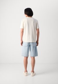 Helles beige Baumwoll-T-Shirt mit kurzen Ärmeln, kombiniert mit hellblauen Denim-Shorts mit zwei Gesäßtaschen und umgeschlagenem Saum. Weiße Sneakers.